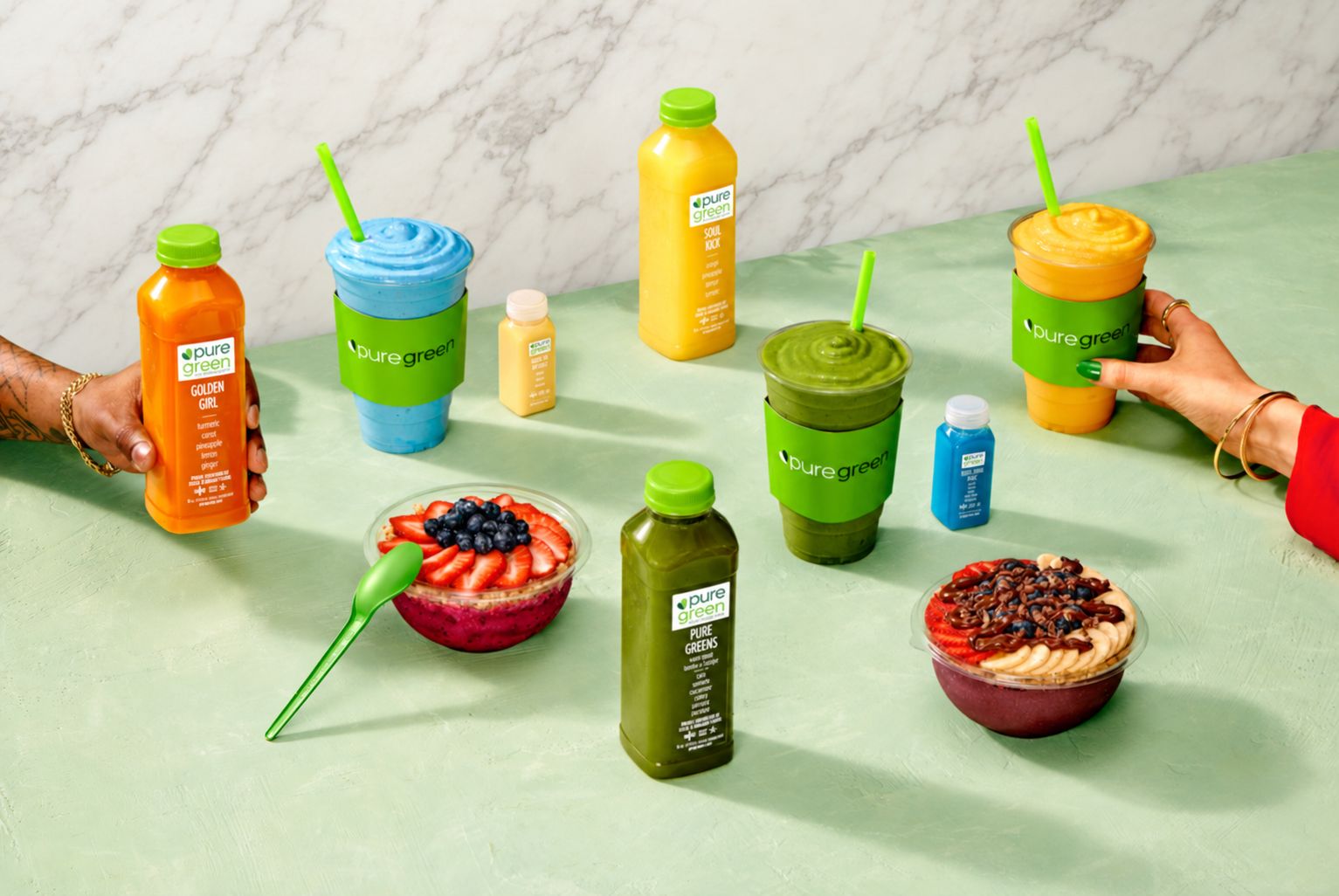 Pure Green Golden Girl and Pure Greens cold-pressed juices, three superfood smoothies, wellness shots and two acai bowls on a mint-green table